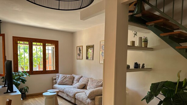 Una sala de estar acogedora con un sofá, una mesita y un televisor. Se ven ventanas con persianas, una escalera y estantes con objetos decorativos.