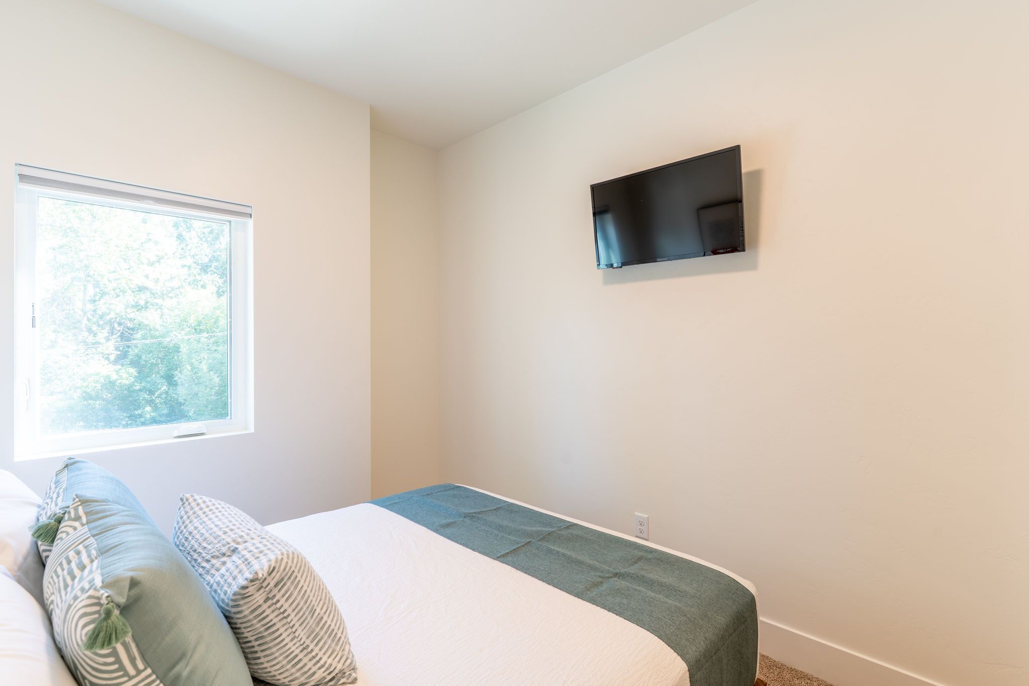 A bedroom with a bed, pillows, a window, and a flat-screen TV mounted on the wall.