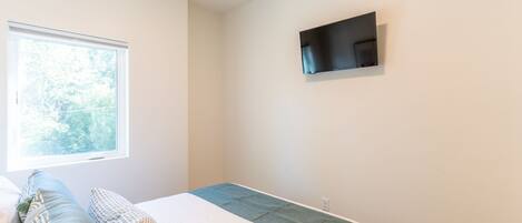 A bedroom with a bed, pillows, a window, and a flat-screen TV mounted on the wall.