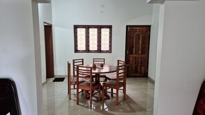 Uma área de refeições com uma mesa redonda de madeira e seis cadeiras, uma janela com grades e uma porta de madeira.