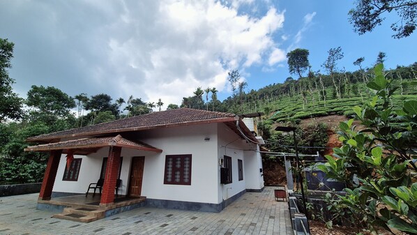 A single-story house with a tiled roof, a porch with a bench, and a view of a lush green hillside.