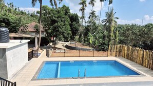 Piscina retangular com duas escadas de metal, cercada por uma vedação de bambu, com vista para uma paisagem verde exuberante e um céu claro.