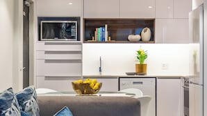 Una cocina moderna con armarios blancos, microondas integrado y lavavajillas. Hay un sofá gris con almohadas azules y un cuenco de limones sobre la mesa.