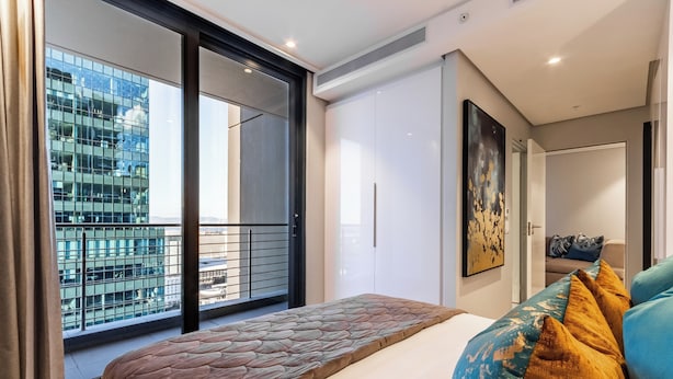 Un dormitorio moderno con una cama grande, vista a un paisaje urbano a través de un ventanal corredero y un balcón con barandilla.