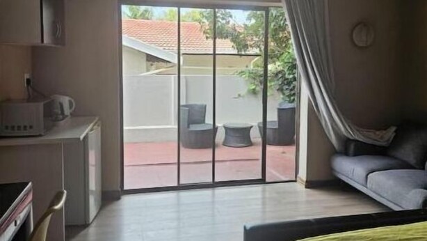 A hotel room with a bed, a desk with a computer, a chair, and a sofa. There is a sliding glass door leading to an outdoor area with chairs and a table.