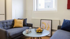 A modern living room with a grey sofa, a small round table with a fruit bowl and a mug, and a framed banana artwork on the wall. - Cranborne House - Campus Accommodation (Bournemouth)