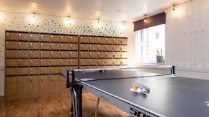 A ping pong table with a net, a wooden cabinet with multiple compartments, and a window with a view of a building. - Cranborne House - Campus Accommodation (Bournemouth)