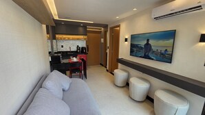A modern hotel room with a sofa, ottomans, a dining area with a red chair, and a large TV on the wall. - Flat vip 02C Bela Vista by La Playa (Ipojuca)