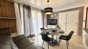 Un salon moderne avec un canapé en cuir, une table à manger avec des chaises, un téléviseur fixé au mur et un coin cuisine visible au fond.