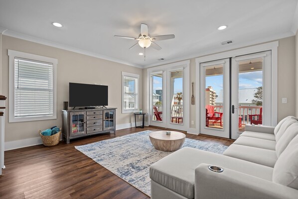 A living room with a ceiling fan, a flat-screen TV, a wooden coffee table, a sofa, and a view through glass doors to an outdoor deck.
