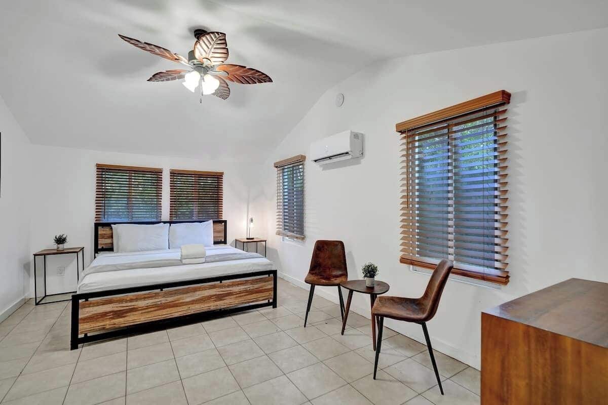A bedroom with a bed, two chairs, a small table, a ceiling fan, and a window with blinds.