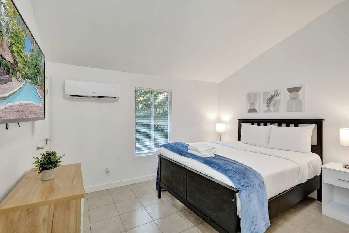 A bedroom with a bed, bedside tables, a wall-mounted air conditioner, a window with blinds, and a small plant on a wooden nightstand.