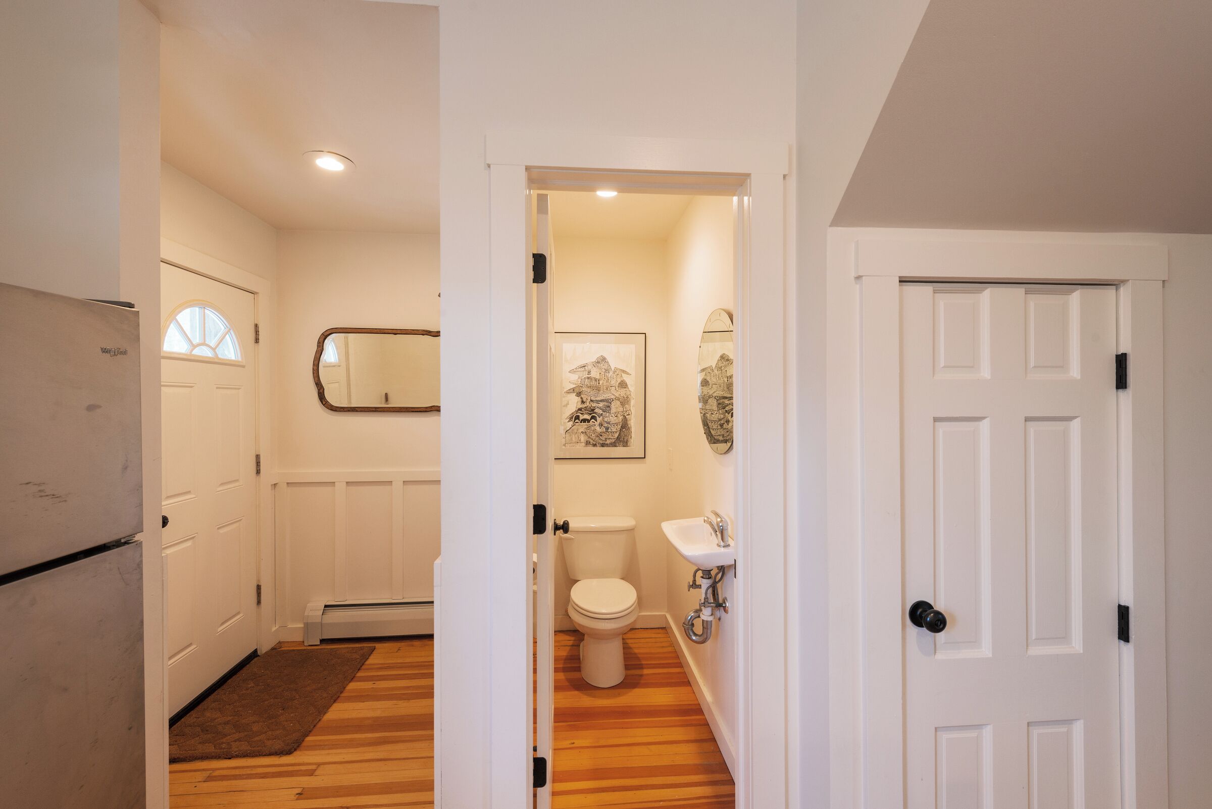 Un couloir étroit mène à une salle de bain équipée de toilettes, d’un lavabo et d’un miroir au mur.