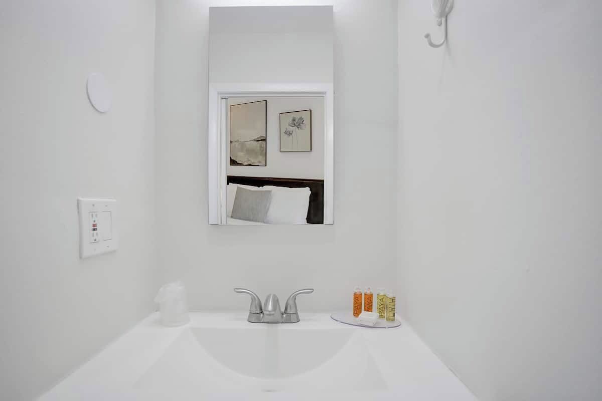Une salle de bain moderne avec un lavabo blanc, un miroir au-dessus et une fenêtre ornée d’une œuvre d’art.