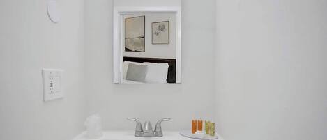 Une salle de bain moderne avec un lavabo blanc, un miroir au-dessus et une fenêtre ornée d’une œuvre d’art.