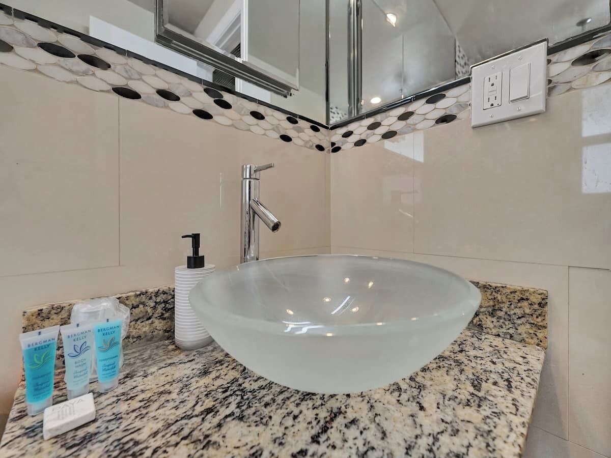 Un baño con un lavabo de vidrio grande, encimera de granito, espejo y artículos de aseo.