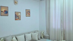 A modern living room with a sofa, three abstract paintings, and a large window with curtains. - Fefe's Mikocheni 1 BR Apartment (Dar es salaam)