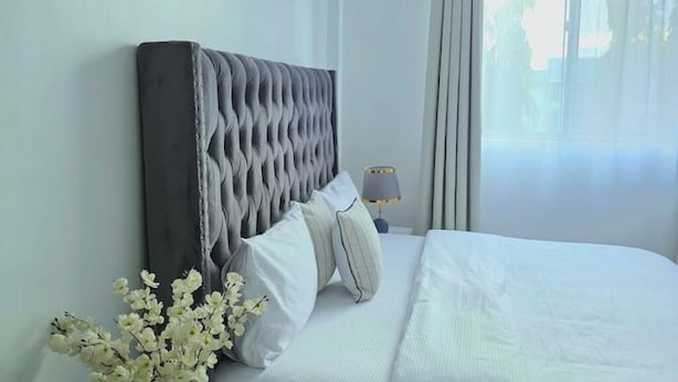 A neatly made bed with white linens and pillows, a tufted headboard, and a bedside table with a lamp. - Fefe's Mikocheni 1 BR Apartment (Dar es salaam)