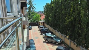A parking area with several cars parked in rows, a building with a balcony on the left, and a tall tree on the right. - Fefe's Mikocheni 1 BR Apartment (Dar es salaam)