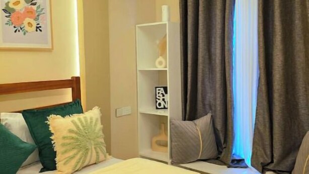 Une chambre d’hôtel comprenant un lit, une table de chevet, une commode, une étagère fixée au mur et une fenêtre avec des rideaux.