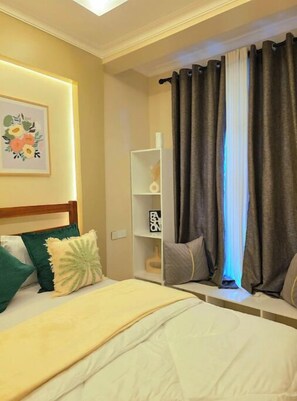 A hotel room with a bed, a bedside table, a nightstand, a wall-mounted shelf, and a window with curtains.