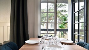Ein Esstisch für zwei Personen, gedeckt mit Tellern, Besteck und Gläsern, platziert in der Nähe eines Fensters mit Blick auf Bäume und Gebäude.