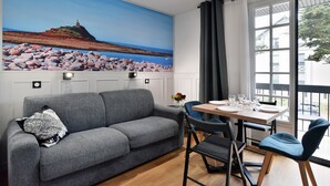 Ein Wohnzimmer mit grauem Sofa, einem Esstisch mit Stühlen und einem großen Gemälde mit Küstenlandschaft.