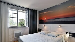 Ein Schlafzimmer mit einem großen Bett, einem Fenster mit Vorhängen, einer Wandlampe und einem Sonnenuntergangsmalerei.