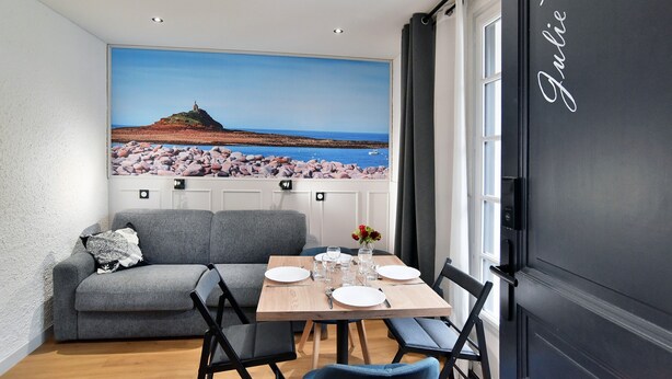 Ein Essbereich mit einem Tisch für zwei Personen, einem grauen Sofa und einem großen Fenster mit schöner Aussicht auf Strand und Meer.