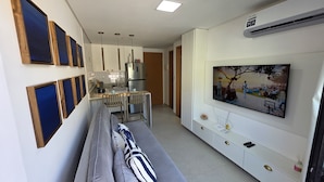 A modern living room with a grey sofa, a dining area with a table and chairs, a kitchen with a refrigerator and a sink, and a mounted TV displaying a beach scene.