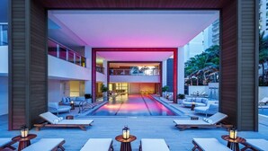 Un vestíbulo de hotel moderno con piscina, sillones de descanso y un balcón en el segundo piso.