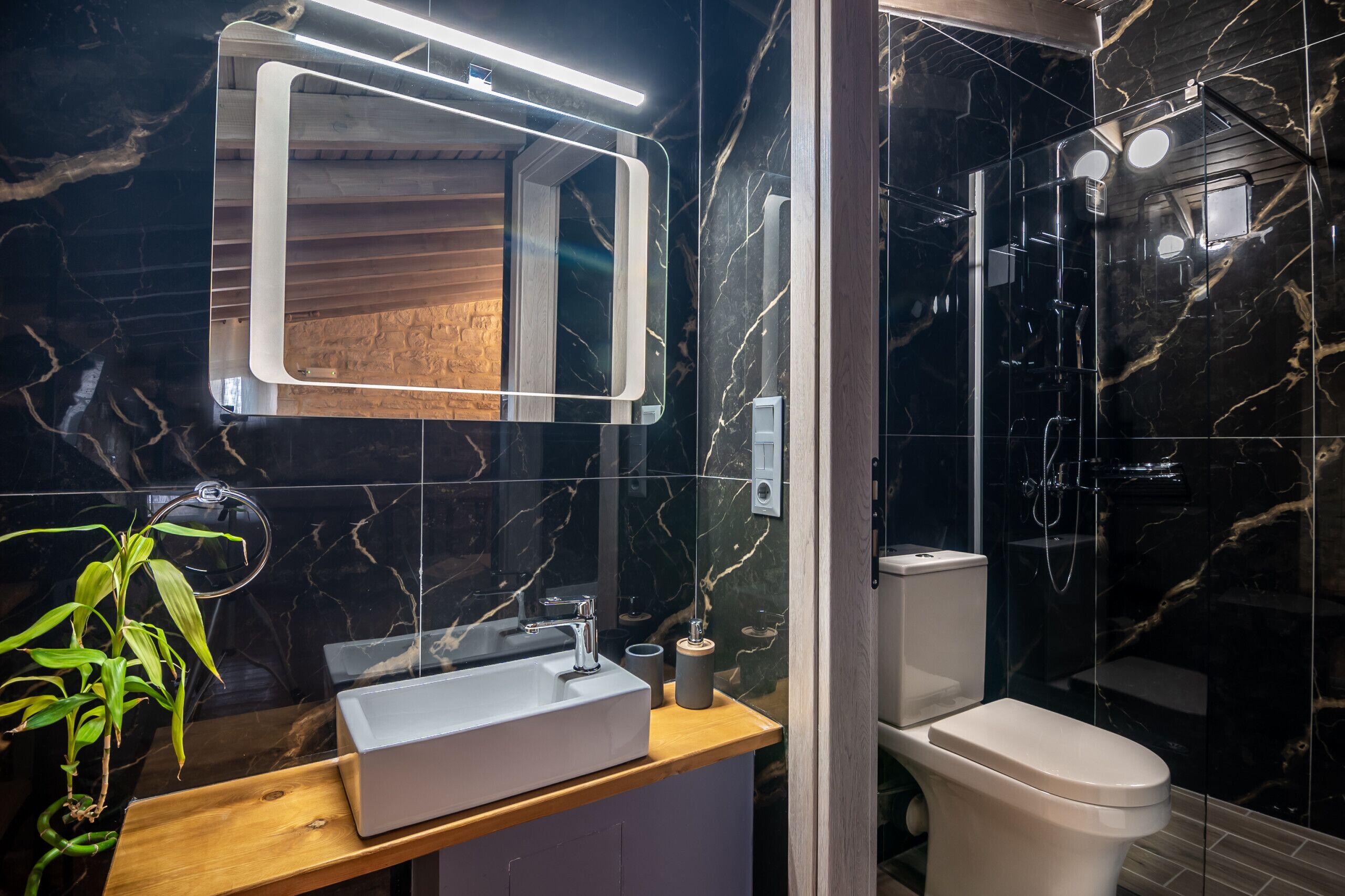 Un baño moderno con detalles de mármol en la pared, un mueble de tocador de madera, un lavabo blanco, un inodoro y una zona de ducha con mampara de cristal.