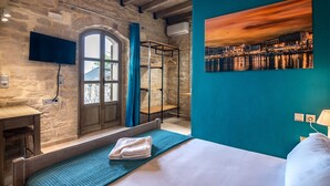 Une chambre d’hôtel avec un mur en pierre, un lit recouvert d’une courtepointe bleue, un téléviseur fixé au mur et un tableau représentant un coucher de soleil sur un paysage urbain.