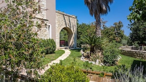 A stone archway leading to a building with a stone facade, surrounded by a well-maintained garden with various plants and a pathway. - Iris Villa (Rethymno)