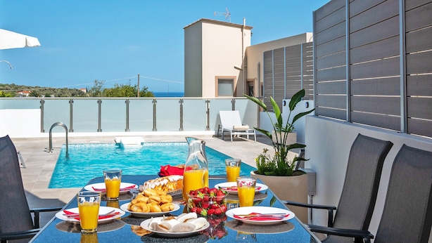 A rooftop pool area with a table set for breakfast, including plates of food, glasses of orange juice, and a pitcher of juice. - Bessie's dream Villa (Rethymno)