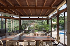 A covered outdoor dining area with a wooden table and wicker chairs, overlooking a pool and lush greenery.