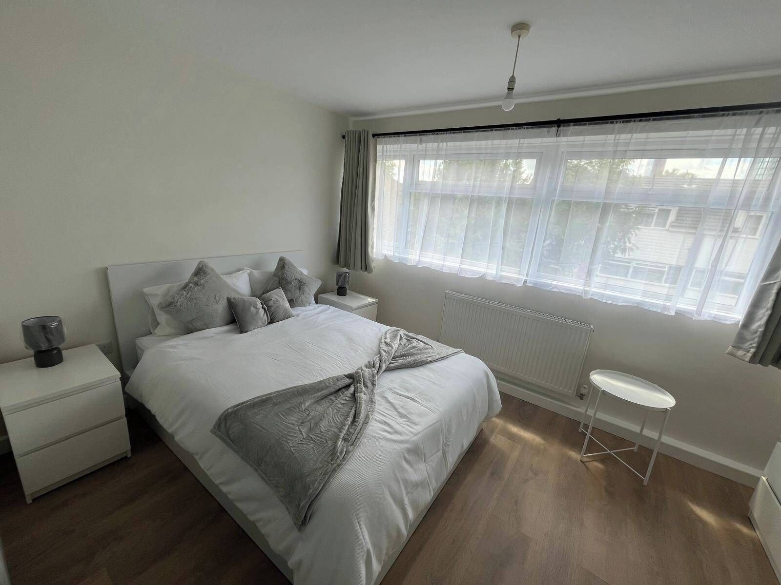 Un dormitorio con una cama, mesitas de noche, una mesilla auxiliar, un radiador y una ventana con cortinas.