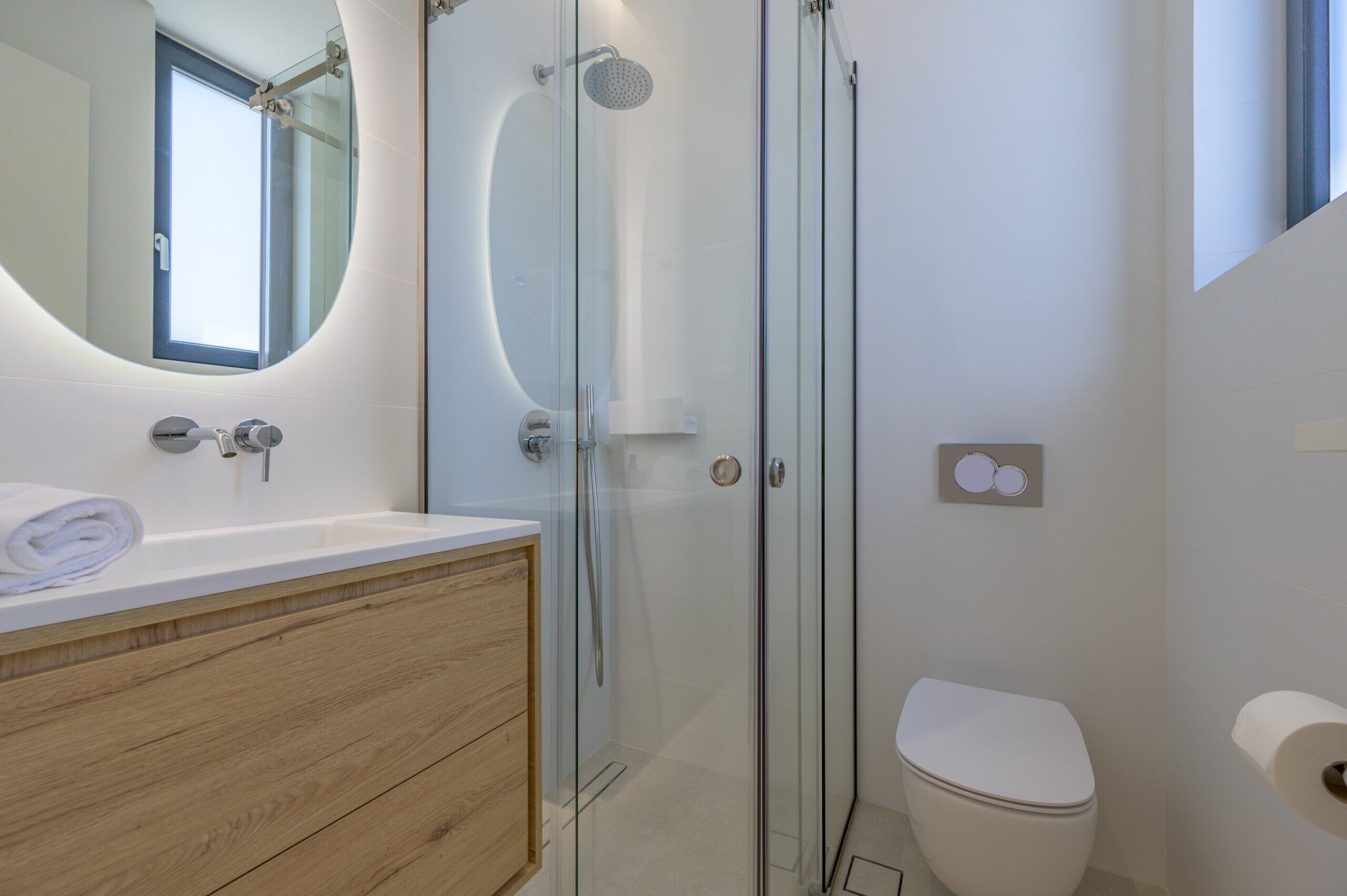 Un baño moderno con ducha envuelta en vidrio, tocador de madera, inodoro y un espejo grande con una ventana arriba.