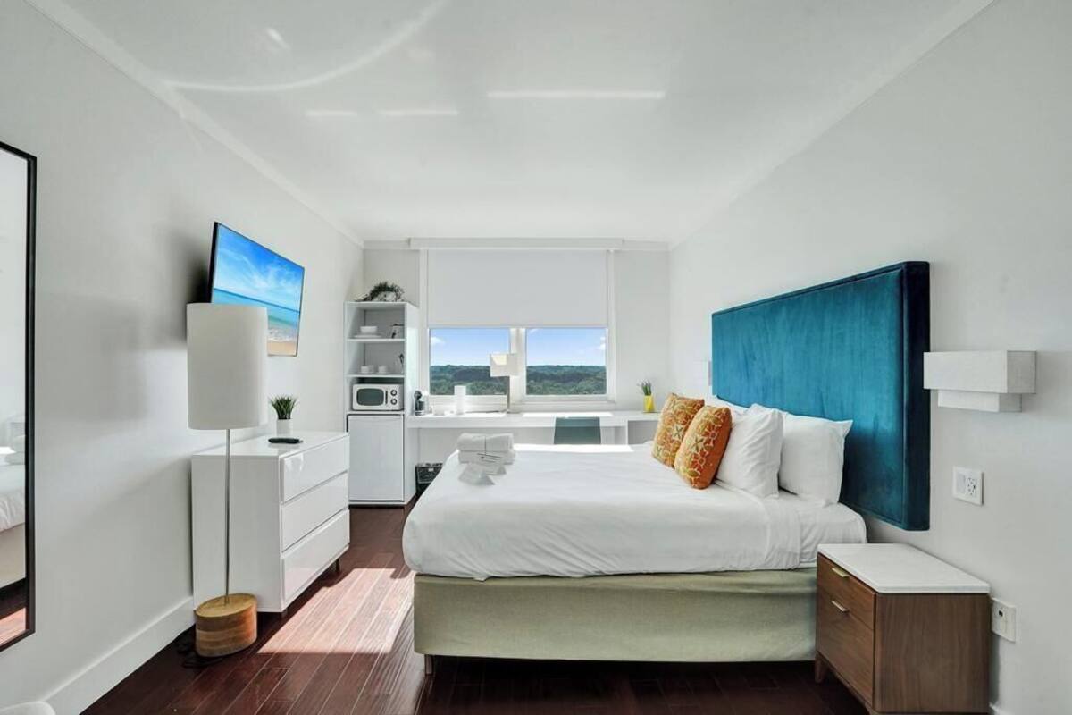 A modern bedroom with a bed, bedside tables, a desk, a chair, a lamp, and a view of the outdoors.