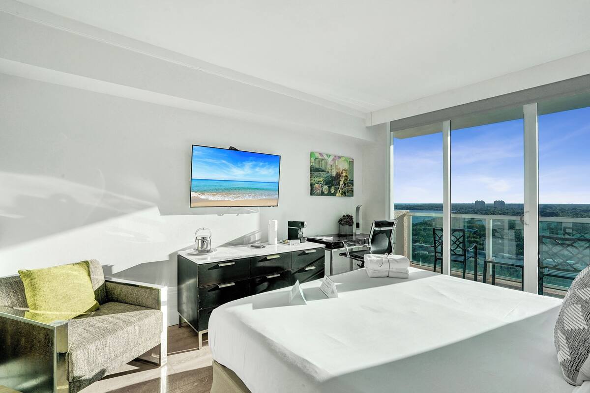 A modern hotel room with a bed, a desk, a chair, a sofa, and a large window offering a city view.