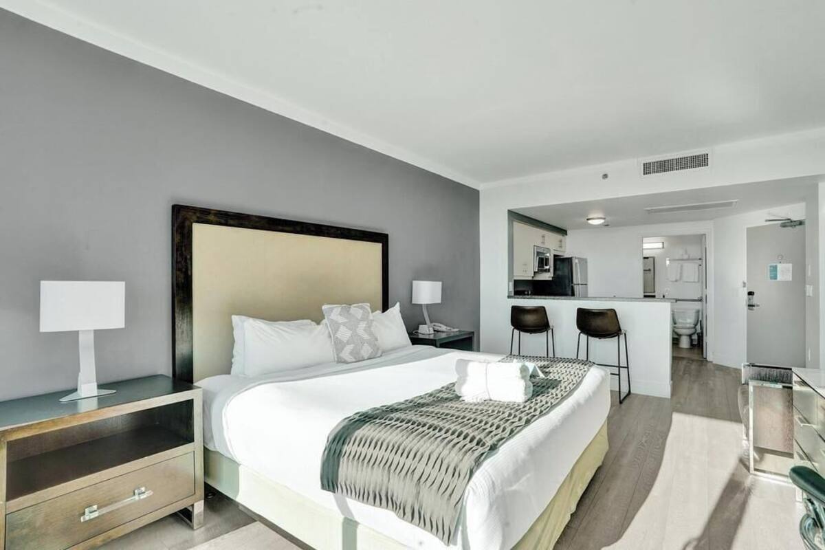 A hotel room with a bed, bedside table, lamp, and a view into the kitchen area.