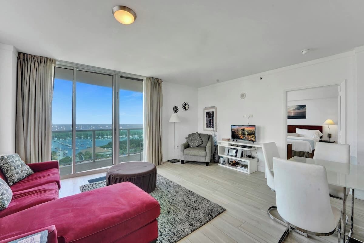 Un salon moderne avec un canapé rouge, un fauteuil blanc et un téléviseur. Une grande fenêtre offre une vue sur la ville.