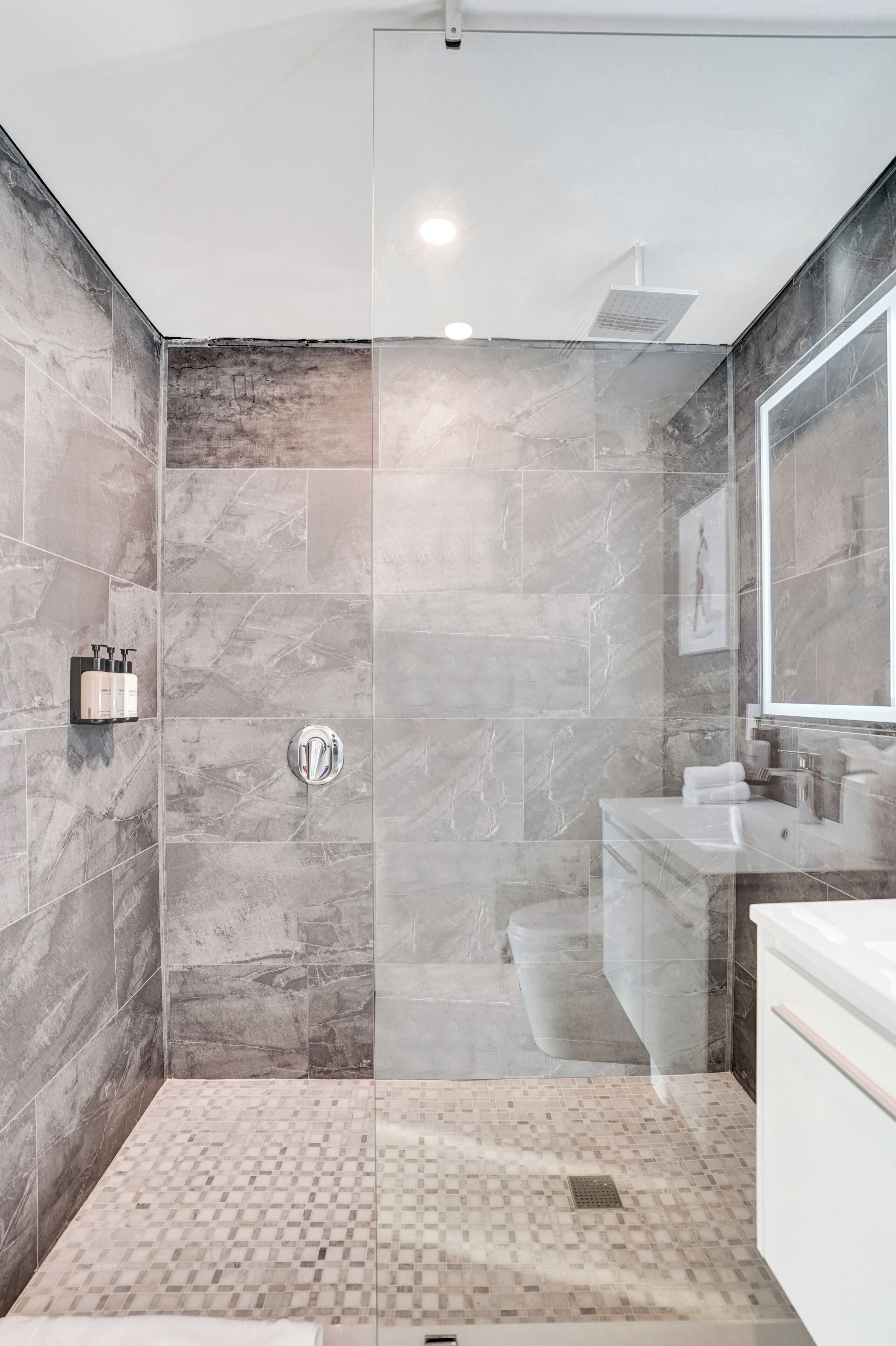 A modern bathroom with a glass shower enclosure, a marble-tiled shower area, a white sink, and a large mirror.