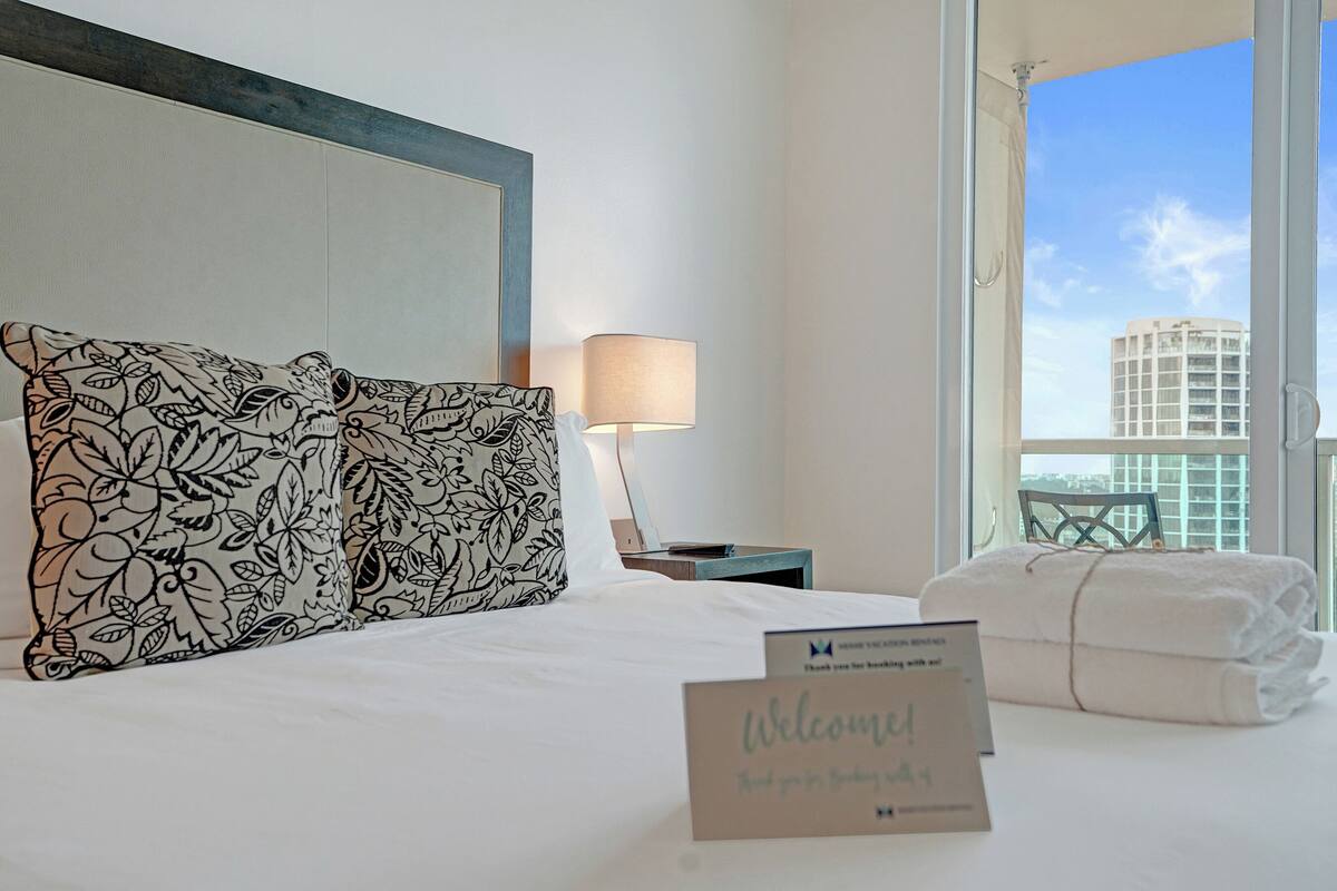A hotel room with a bed, a nightstand, a lamp, a window with a city view, and a welcome sign.