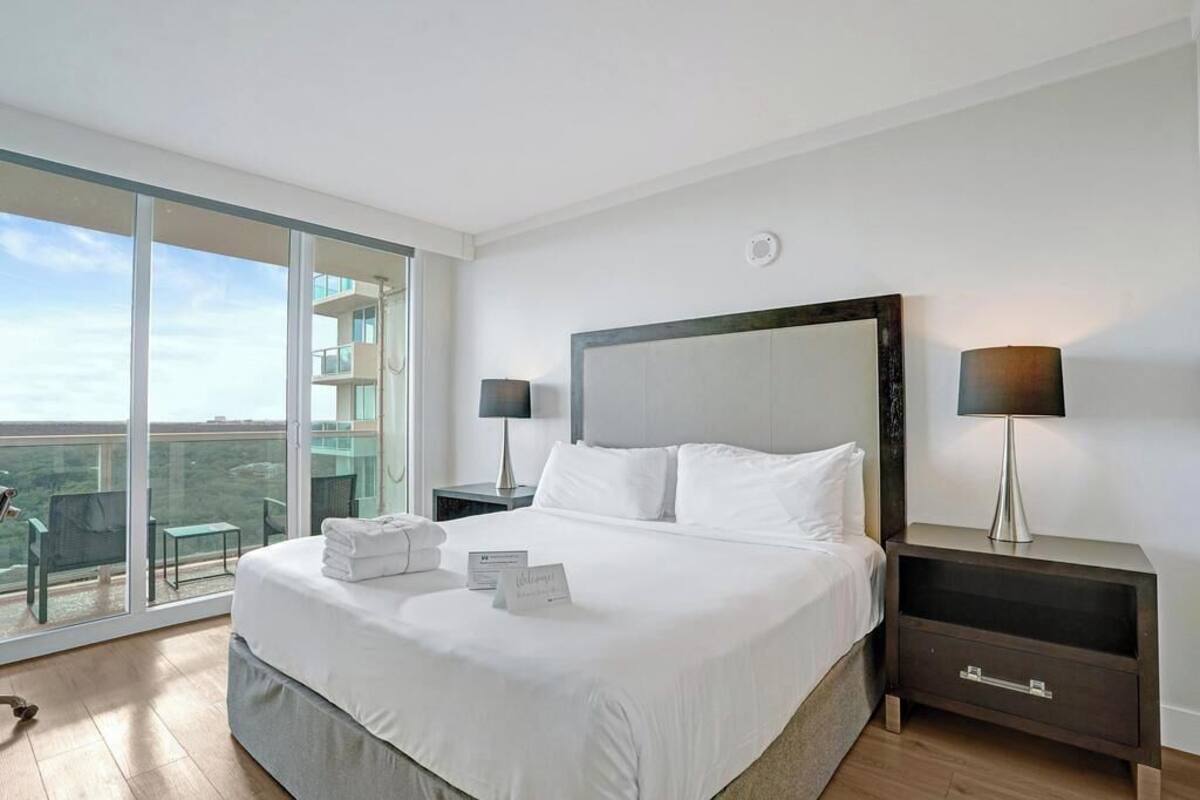 A hotel room with a large bed, two bedside tables, a nightstand, a balcony with a chair, and a view of the city.