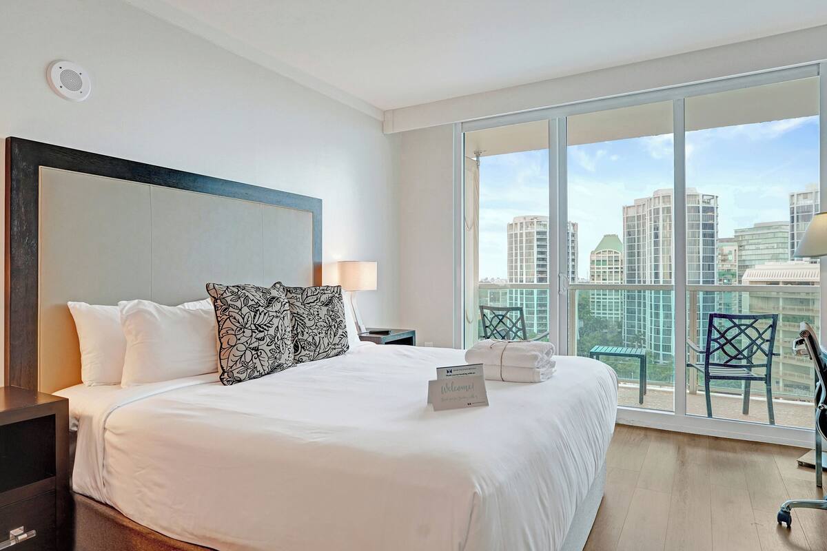 A hotel room with a large bed, a bedside table, a chair, and a balcony with a view of the city.
