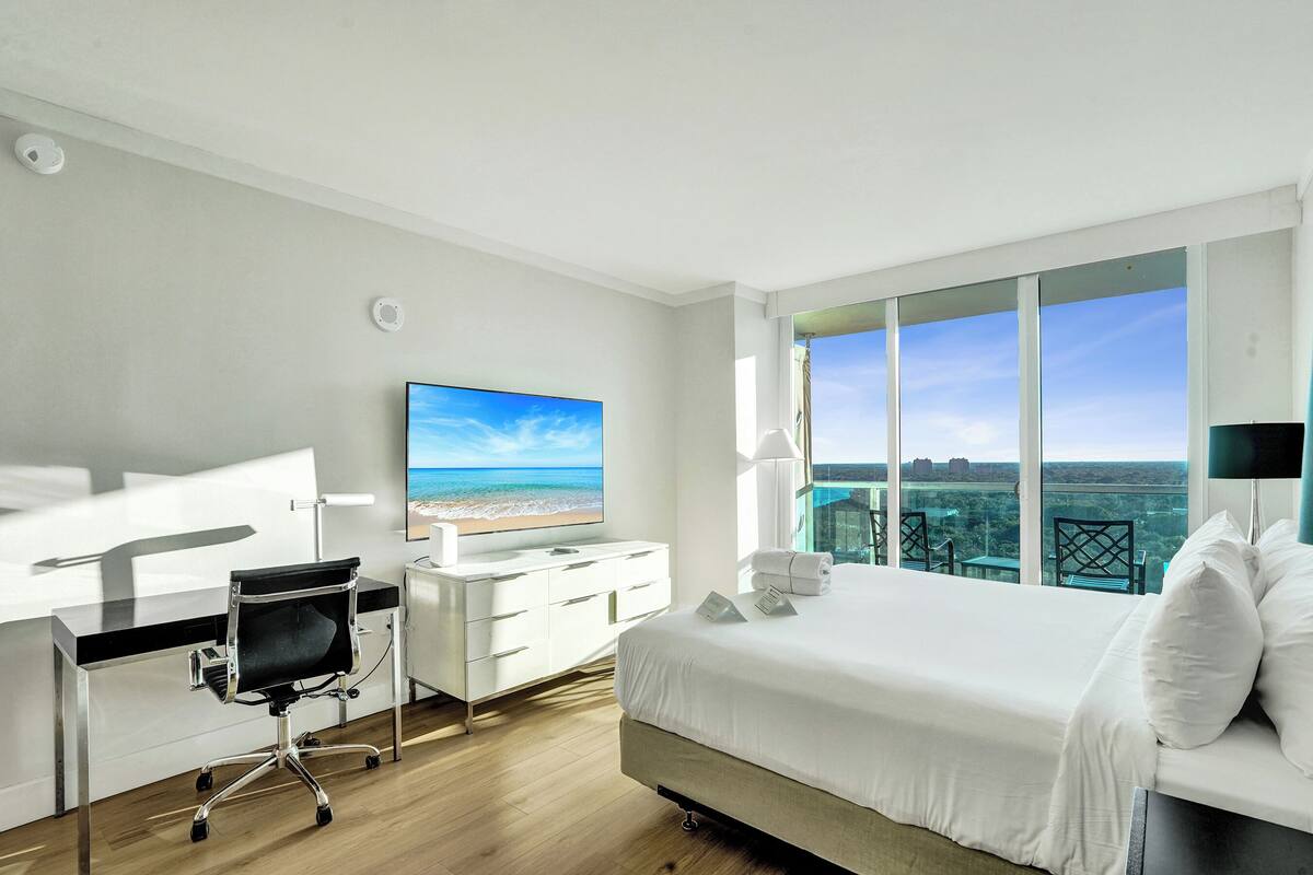 A hotel room with a bed, a desk with a chair, a TV, and a balcony with a view.