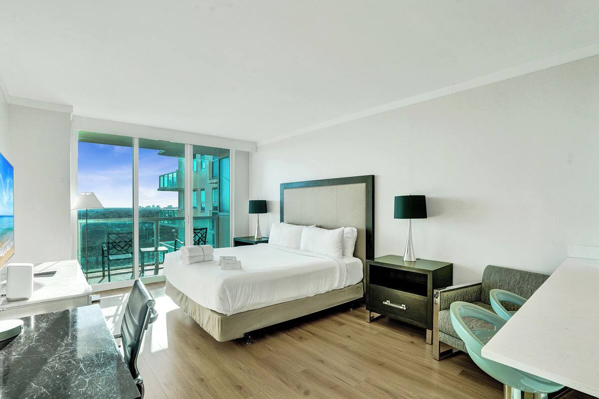 A hotel room with a bed, a desk, a chair, a nightstand, and a view of the ocean.