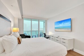 A hotel room with a large bed, a desk, a chair, a lamp, a painting of a beach, and a view of buildings through the window. - Stay Hospitality - Coconut Grove (Miami)