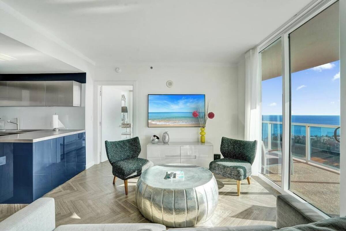 A modern living room with a round coffee table, two patterned armchairs, a flat-screen TV, and a view of the ocean.